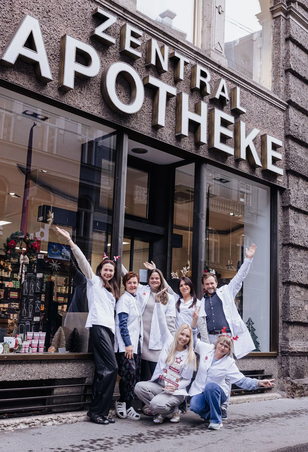 Fröhliches Team mit erhobenen Armen vor dem Schaufenster und dem Schriftzug „Zentral Apotheke“